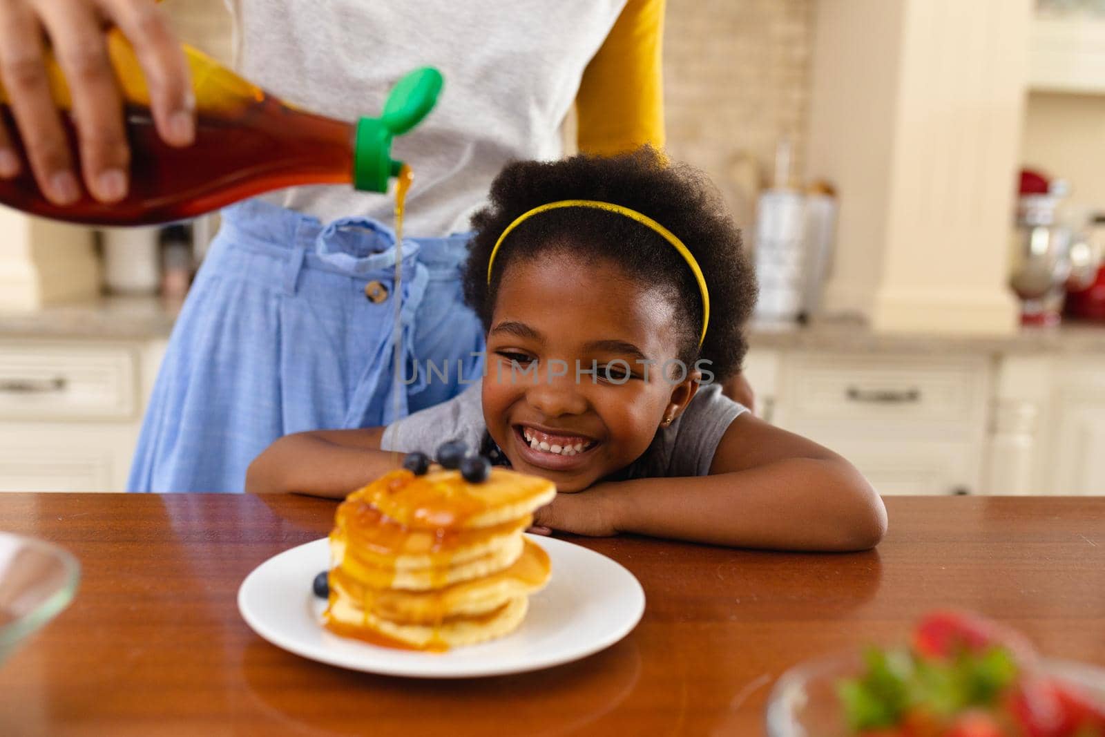 Midsection of mother putting maple syrup on pancake of her smiling african american daughter at home. family, love and togetherness concept, unaltered.