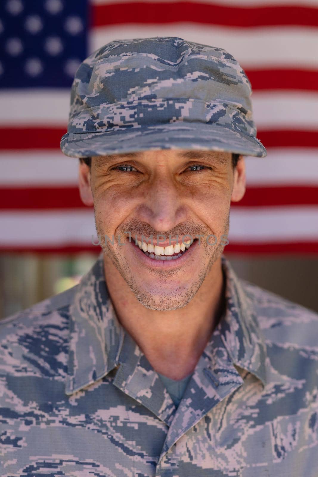 Portrait of smiling caucasian army soldier wearing cap and camouflage uniform against usa flag. people, patriotism and identity concept, unaltered.