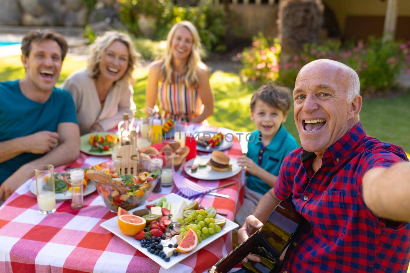 Portrait of senior caucasian man taking selfie with family sitting together at table in garden. family, togetherness and weekend lifestyle concept, unaltered.
