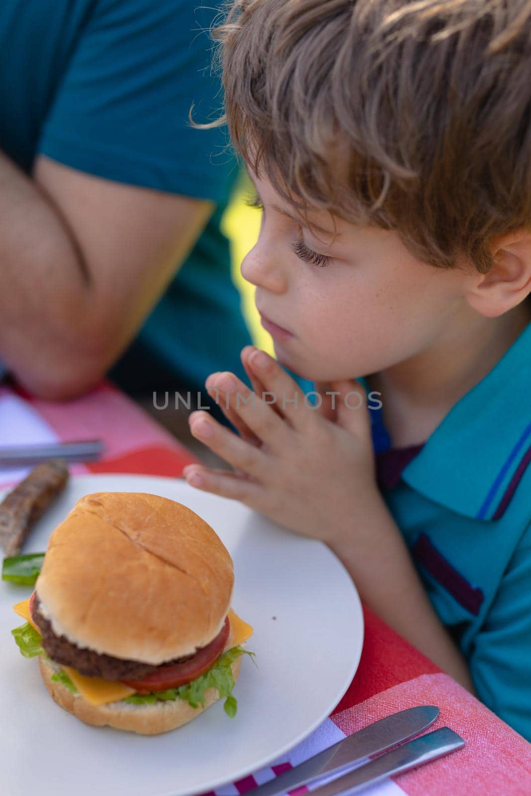 Caucasian boy with hands clasped and eyes closed praying by burger on table in garden. people, food and spirituality concept, unaltered.