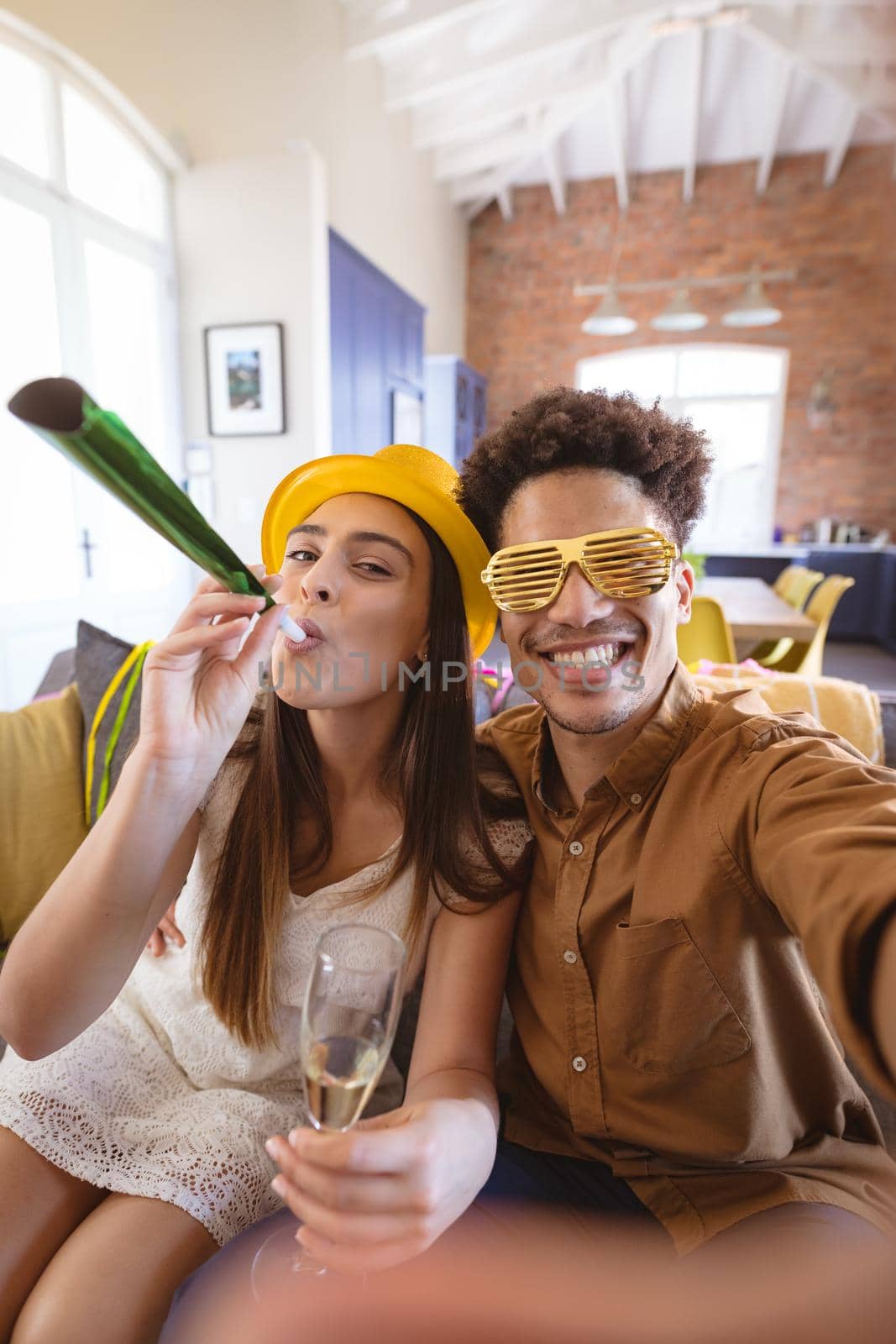 Happy man taking selfie with girlfriend blowing party horn celebrating at home. biracial couple, enjoying and lifestyle.