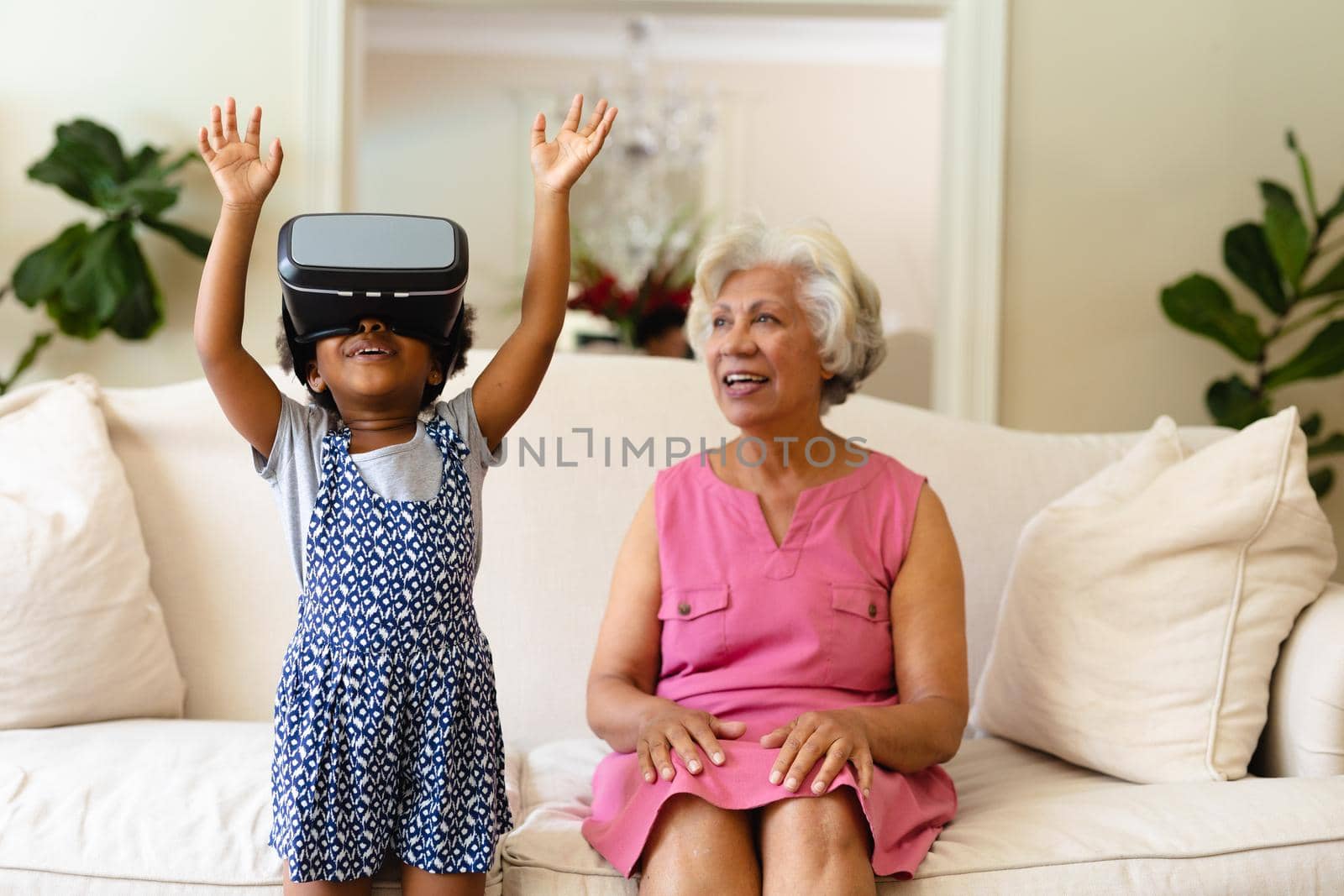 African american grandmother looking at her granddaughter wearing vr headset at home. virtual reality and futuristic technology concept, unaltered.
