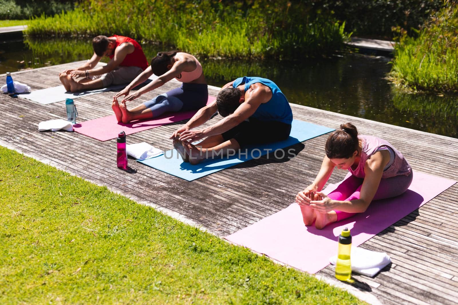 Men and women touching toes while practicing yoga on floorboard by pond in public park by Wavebreakmedia