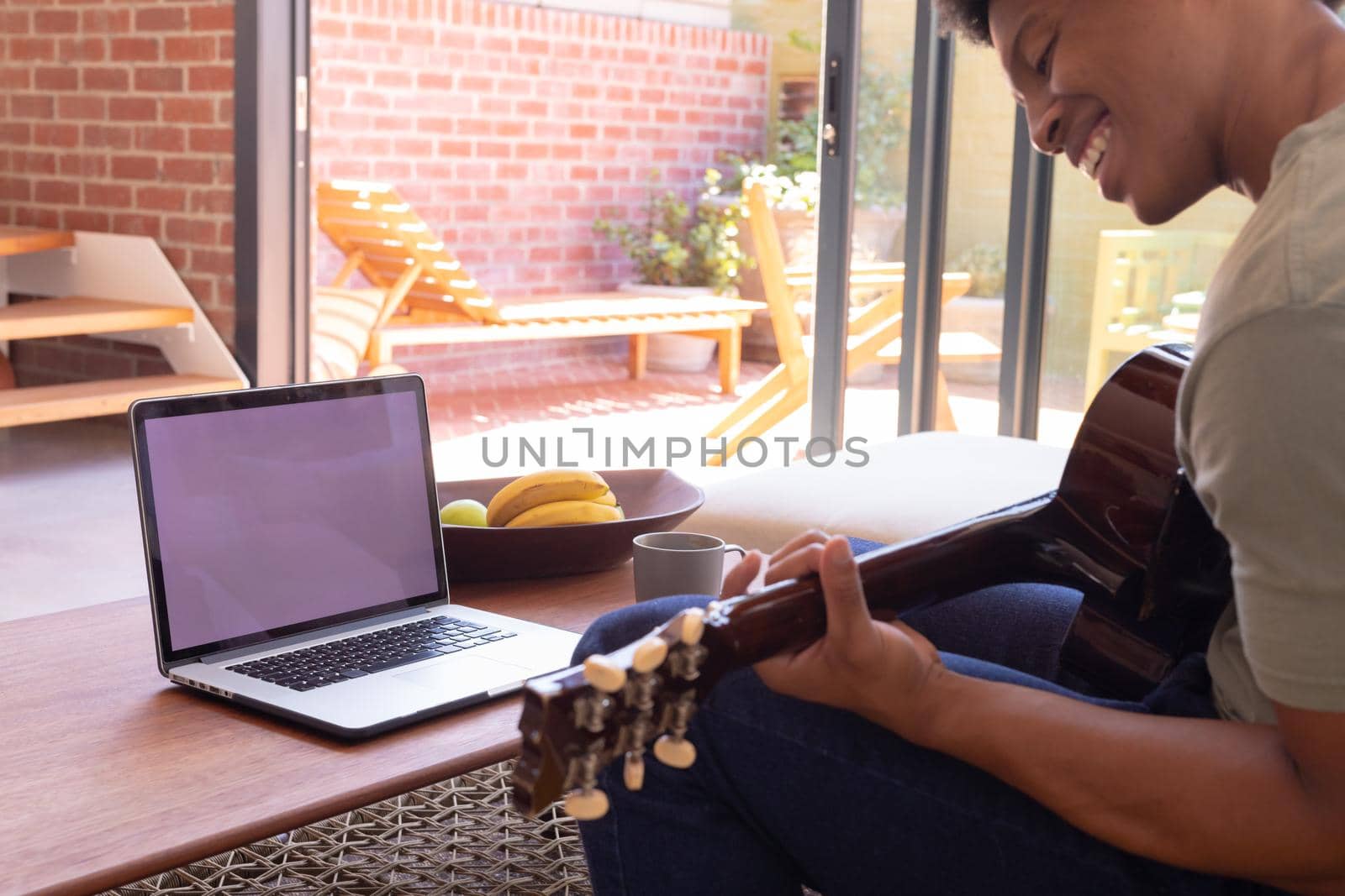 Smiling african american young man playing guitar while using laptop with blank space, copy space. unaltered, lifestyle, leisure activity, music, food, wireless technology and domestic life concept.