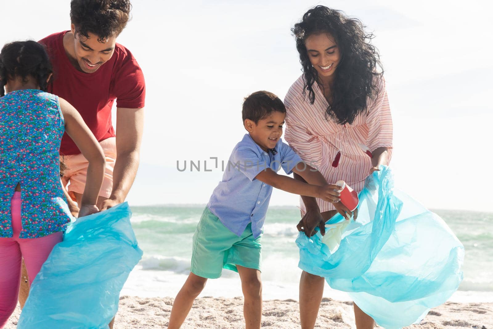 Happy multiracial parents and children collecting plastic waste in large bags at beach on sunny day. lifestyle and environmentalism.