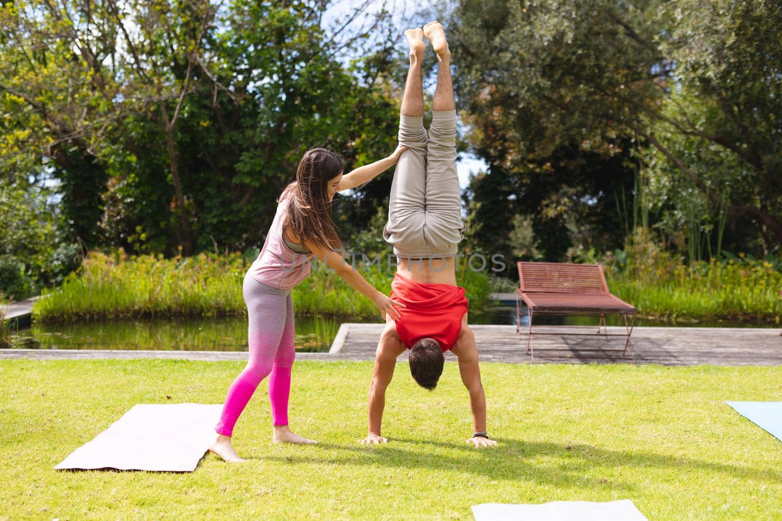 Full length of female instructor assisting man in practicing handstand in park on sunny day. yoga, healthy lifestyle and body care.