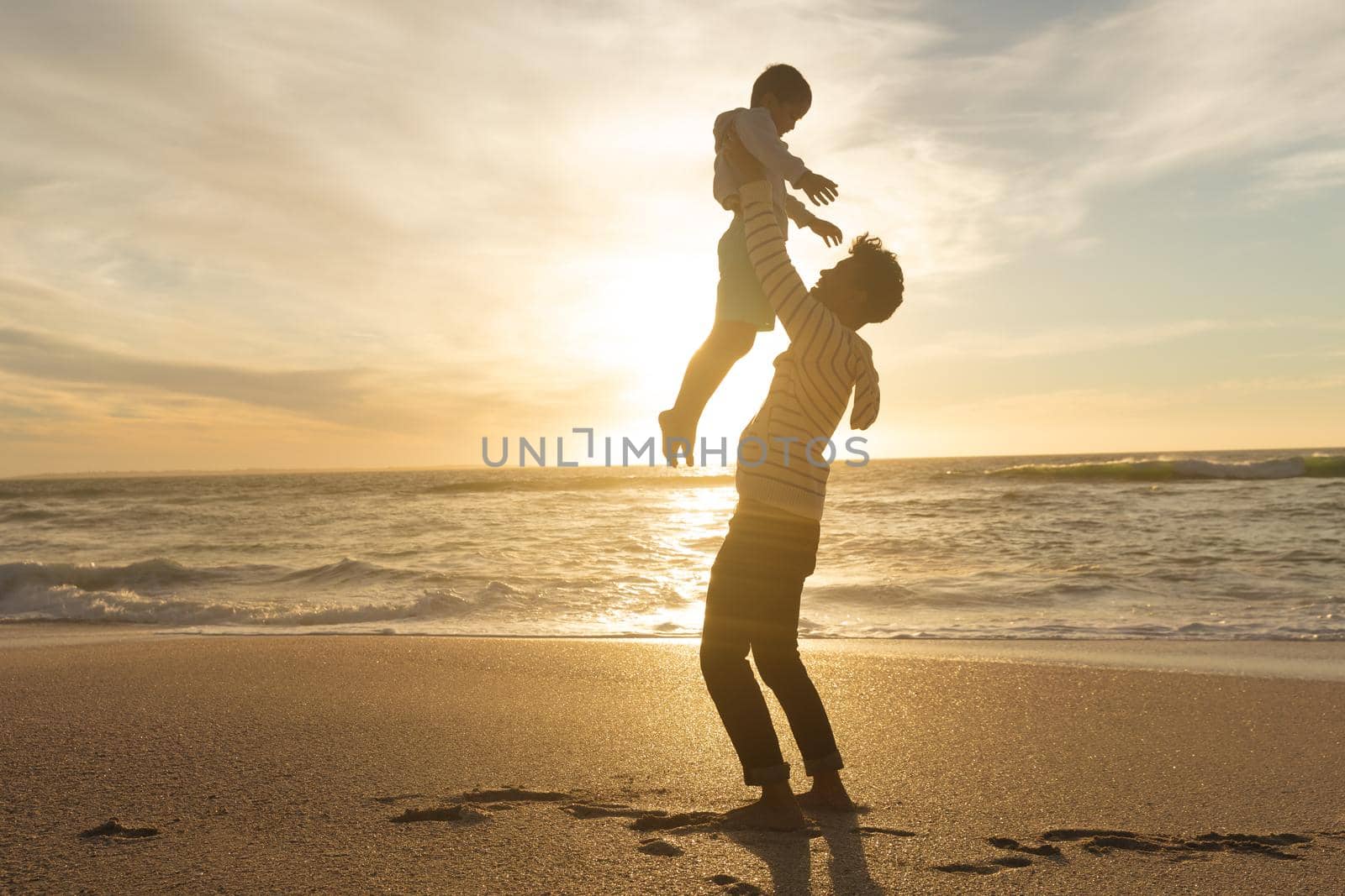 Side view of playful biracial father holding son aloft on shore at beach against sky during sunset. family, lifestyle and weekend.
