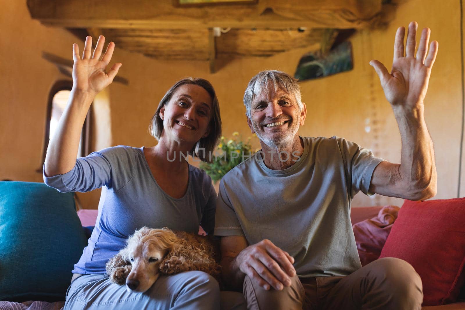 Portrait of mature couple with dog waving hands while sitting on couch in cob home. togetherness, lifestyle and sustainable living.