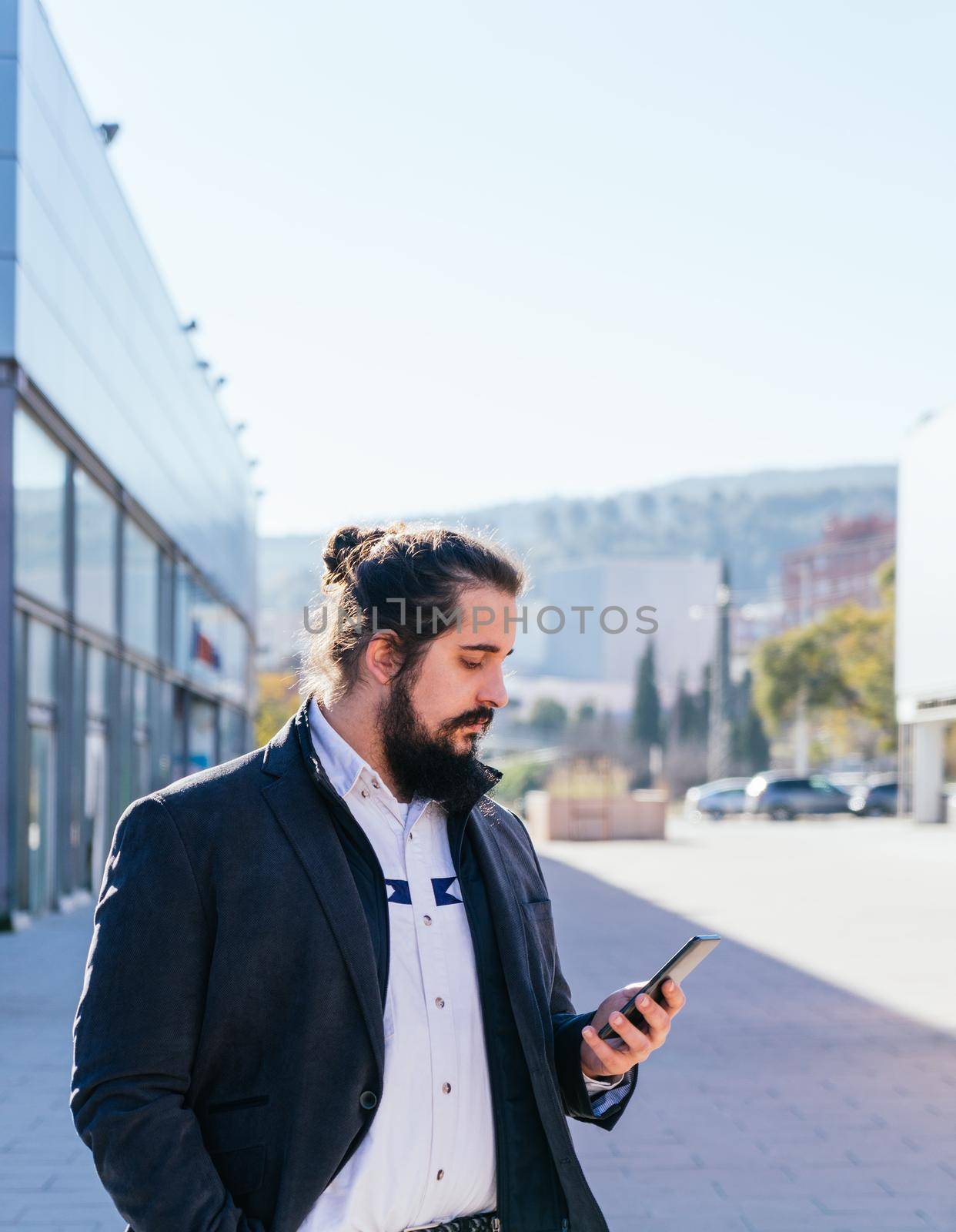 Young businessman with long hair and beard looking smartphone during his work break, on the street next to the offices. Vertical photo on a sunny and clear day.