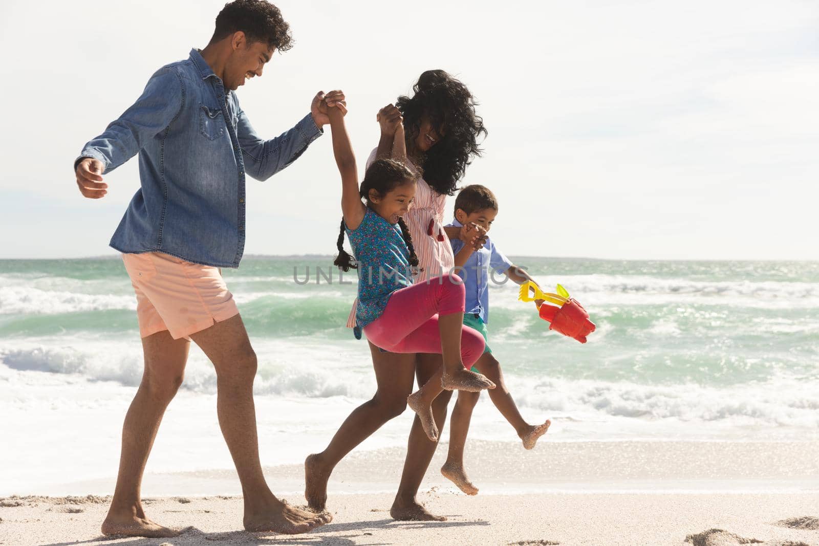 Playful multiracial family walking while holding hands on shore at beach enjoying sunny day by Wavebreakmedia