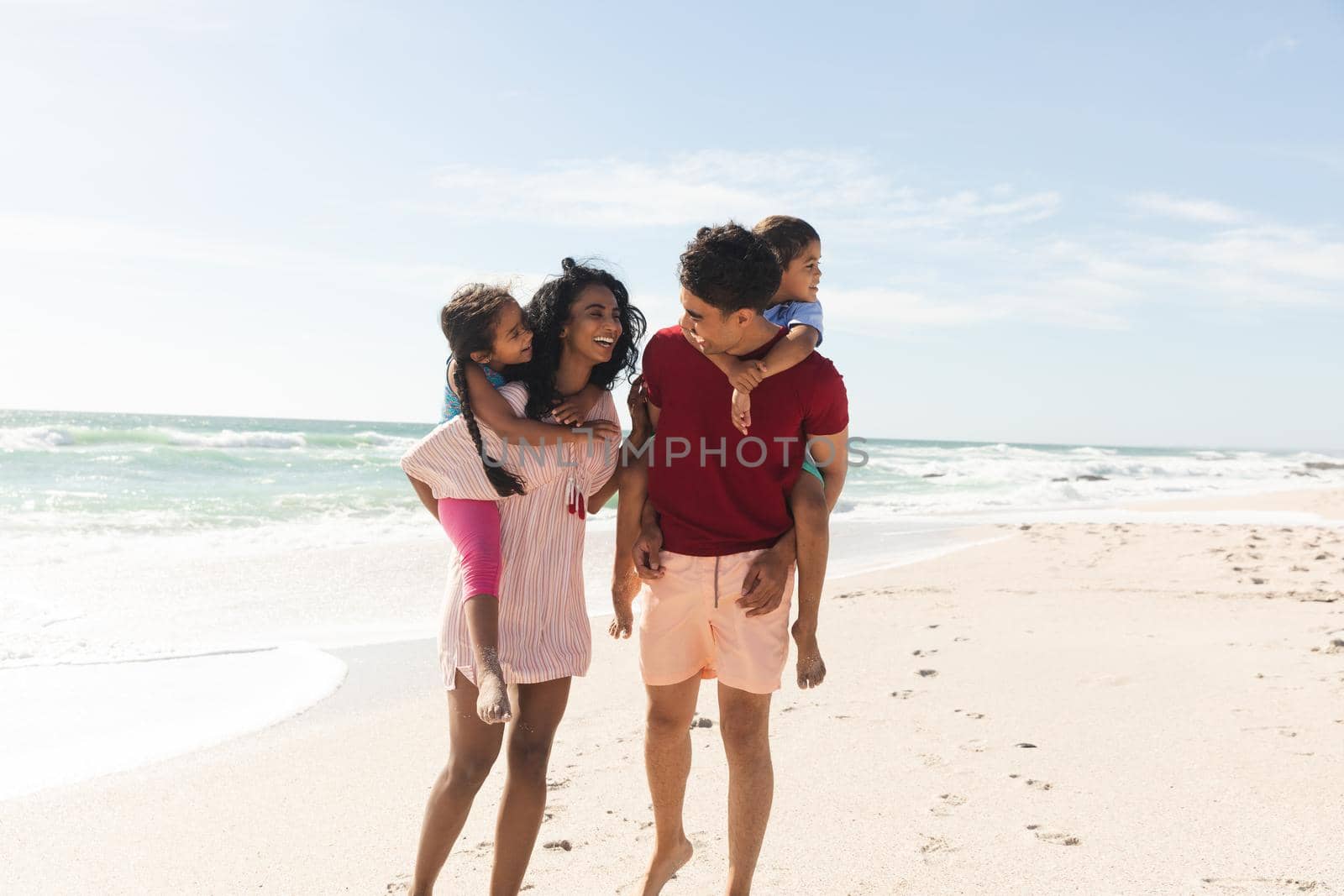 Happy multiracial parents giving piggyback rides to children at beach against sky on sunny day. family, lifestyle and weekend.