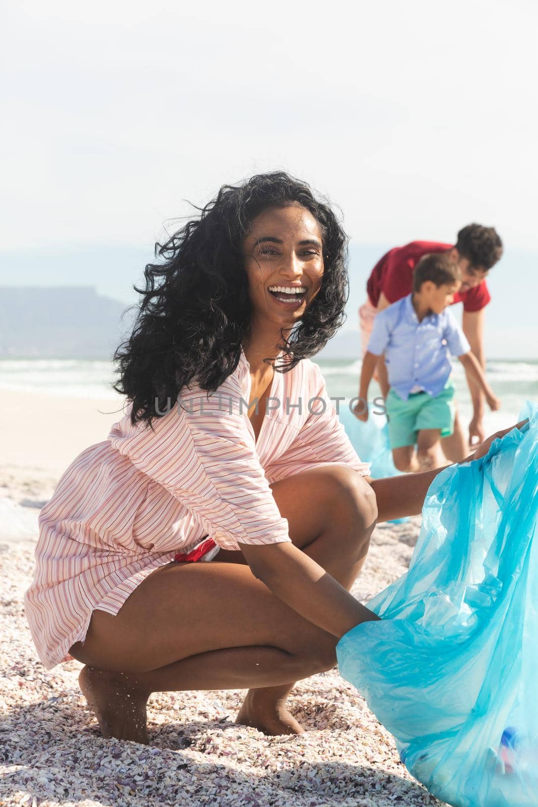 Portrait of cheerful biracial woman collecting waste in blue plastic bag with family at beach. lifestyle and environmentalism.