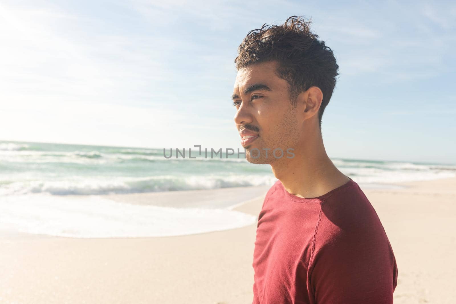 Biracial man looking away while standing at beach against sky on sunny day by Wavebreakmedia