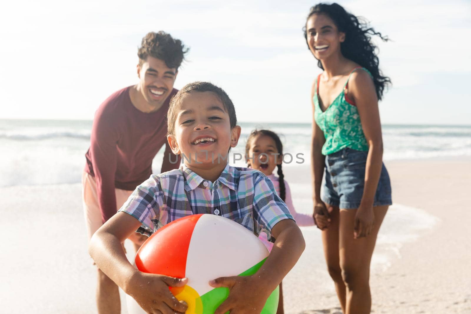 Portrait of smiling biracial boy holding ball with family at beach on sunny day by Wavebreakmedia
