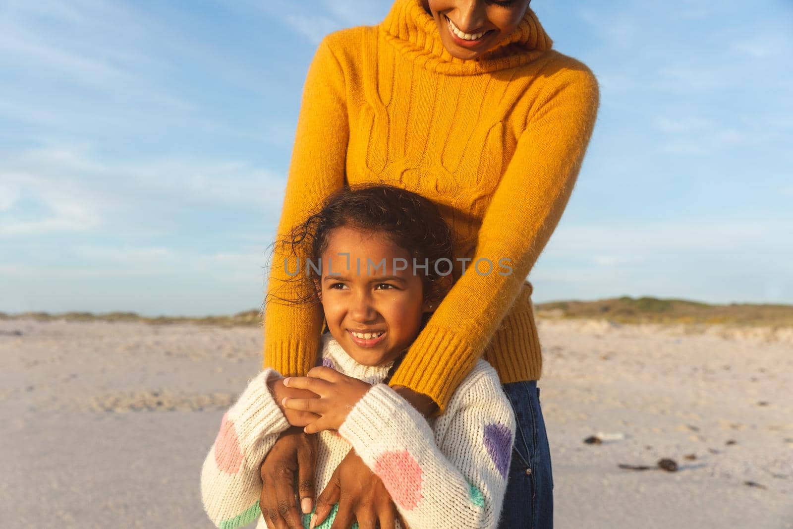 Smiling young biracial woman standing behind daughter looking away at beach against sky. family, lifestyle and weekend.