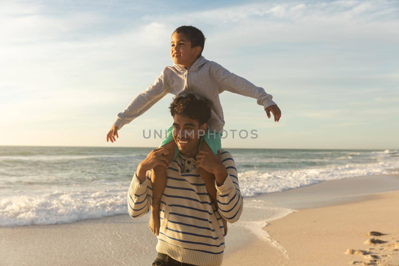 Happy biracial father carrying son on shoulders with arms outstretched enjoying sunset at beach. family, lifestyle and weekend.