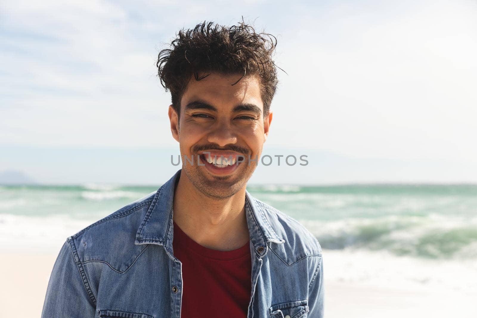 Portrait of smiling biracial man wearing denim shirt at beach on sunny day. lifestyle and weekend.