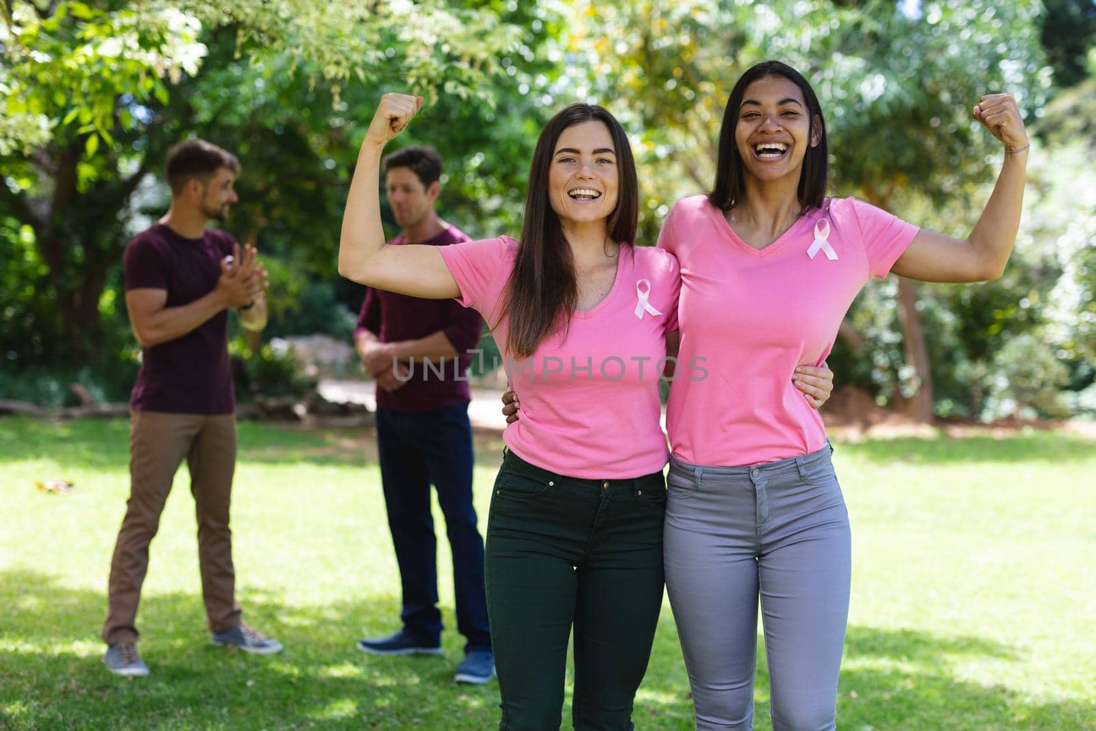 Happy multiracial female friends wearing breast cancer awareness ribbons flexing muscles in park by Wavebreakmedia