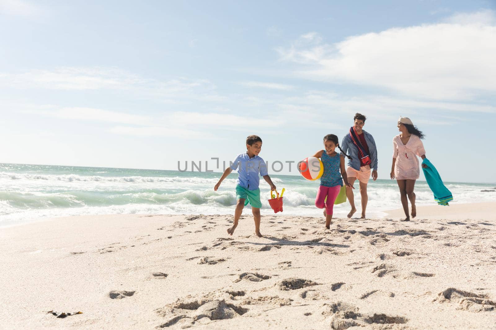 Multiracial parents walking behind children running on sand at beach during sunny day by Wavebreakmedia