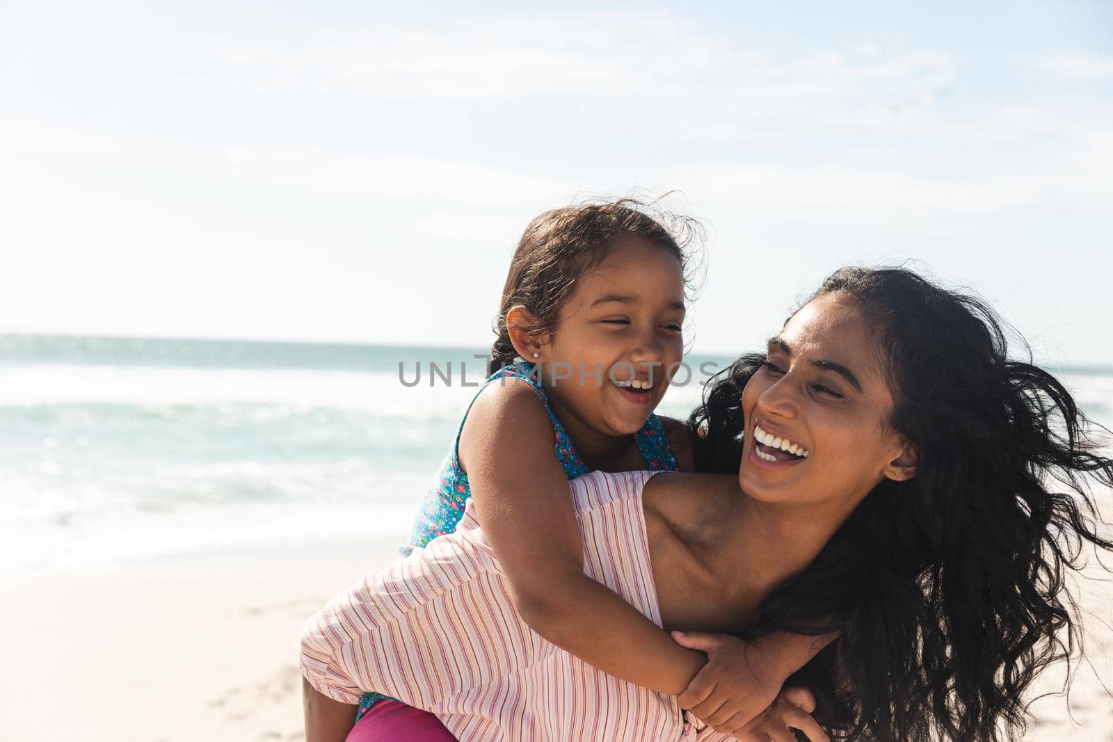 Cheerful biracial woman giving piggyback ride to daughter at beach against sky with copy space. family, lifestyle and weekend.