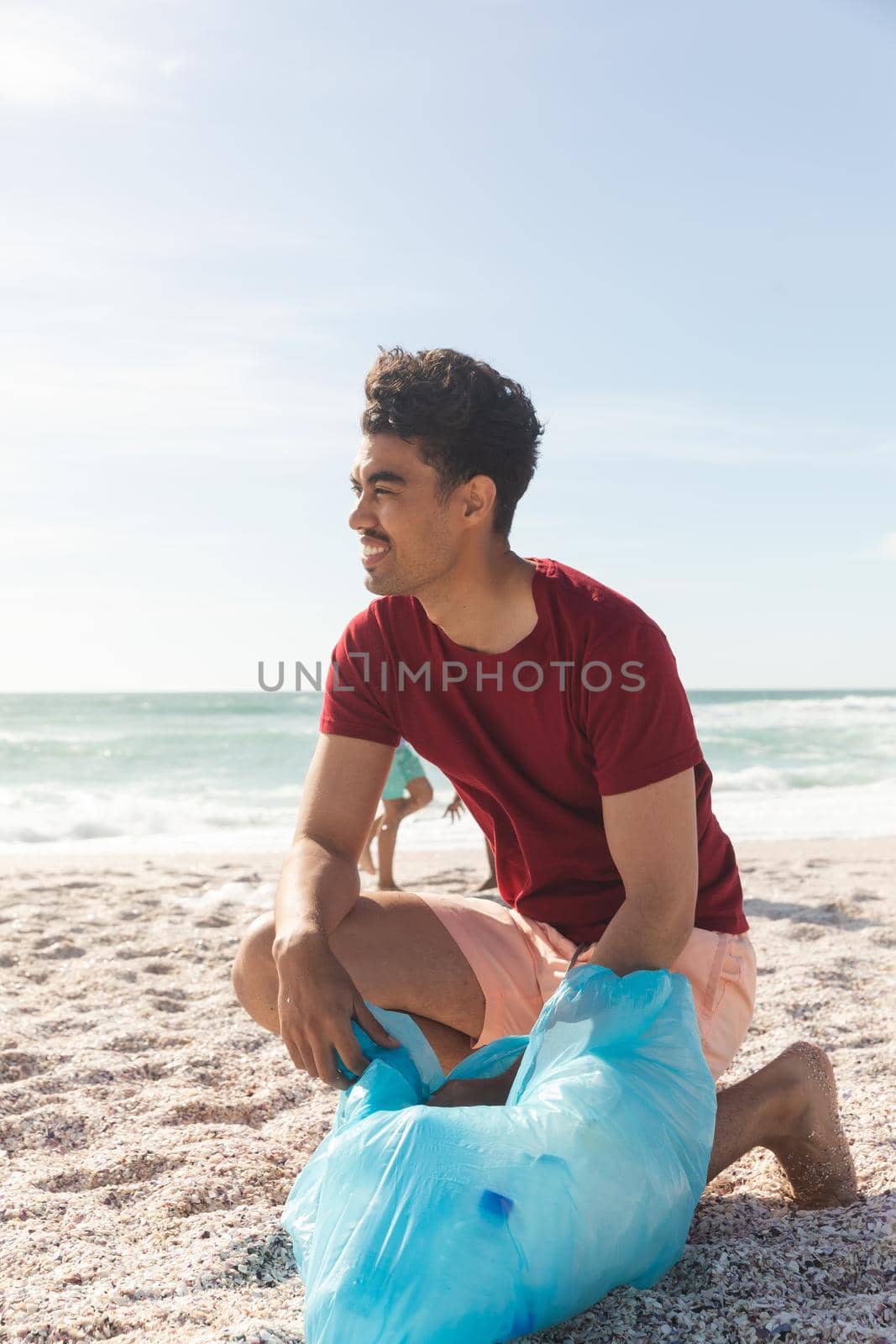 Full length of biracial man collecting garbage in bag at beach against sky with copy space. lifestyle and environmentalism.