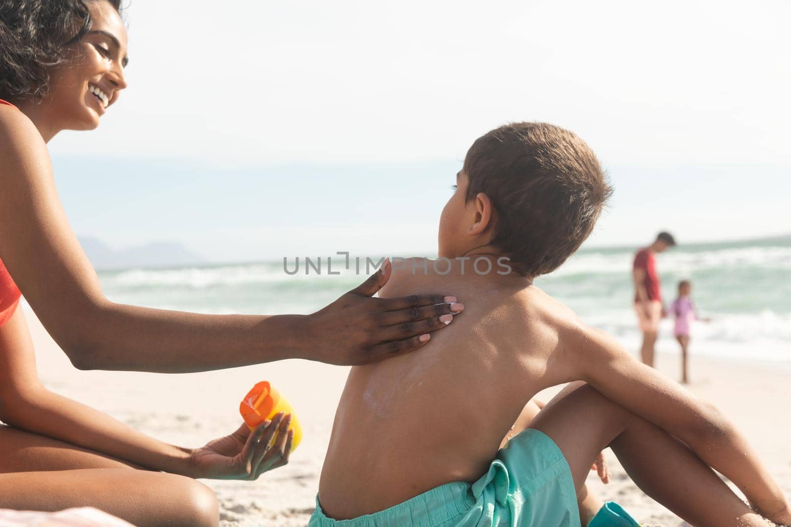 Smiling biracial woman applying sunscreen lotion on shirtless son's back at beach during sunny day. family, lifestyle and care.