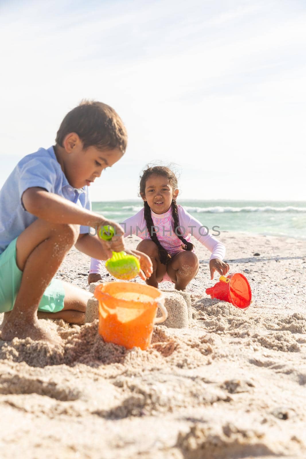 Biracial brother and sister collecting sand in buckets while playing at beach on sunny day by Wavebreakmedia