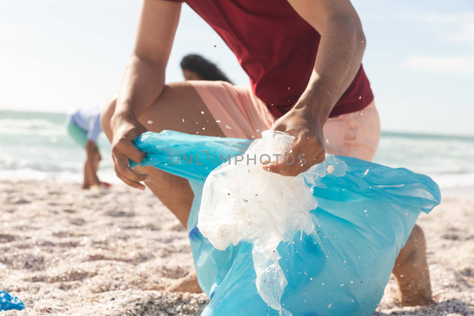 Low section of biracial man collecting plastic waste in bag at beach on sunny day. lifestyle and environmentalism.