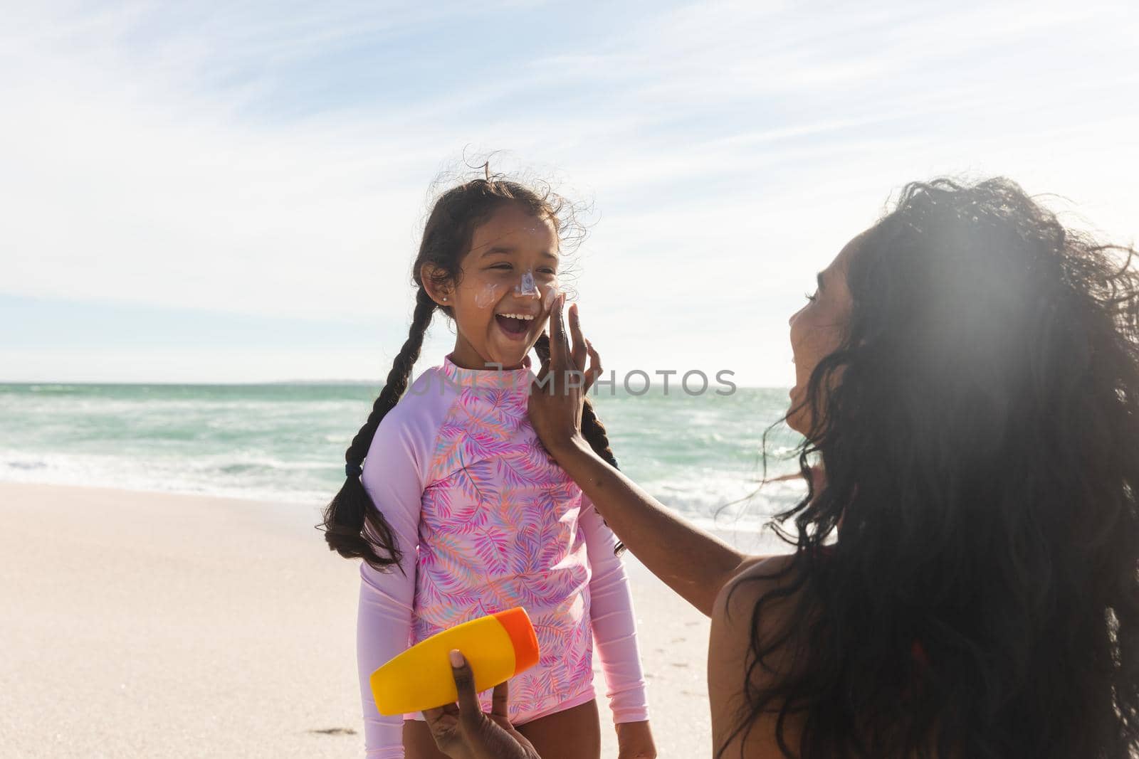 Cheerful biracial girl looking at mother applying sunblock lotion on her nose during sunny day. family and lifestyle and care.