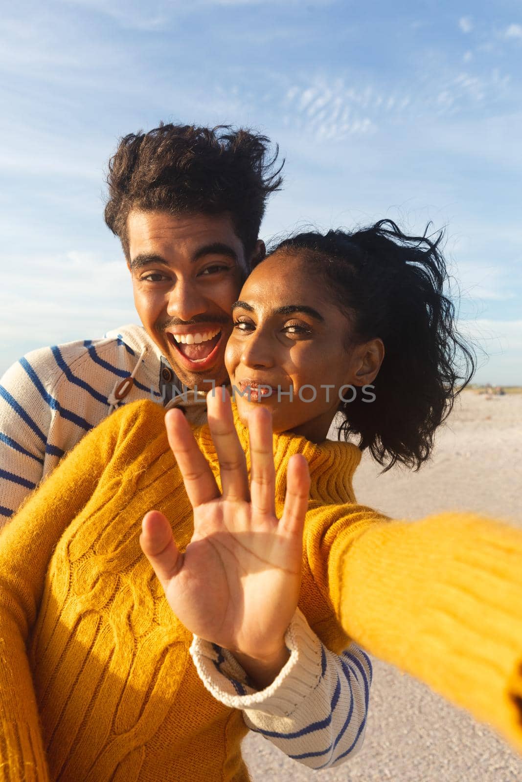 Portrait of smiling biracial woman taking selfie with boyfriend gesturing stop sign at beach. lifestyle, love and weekend.