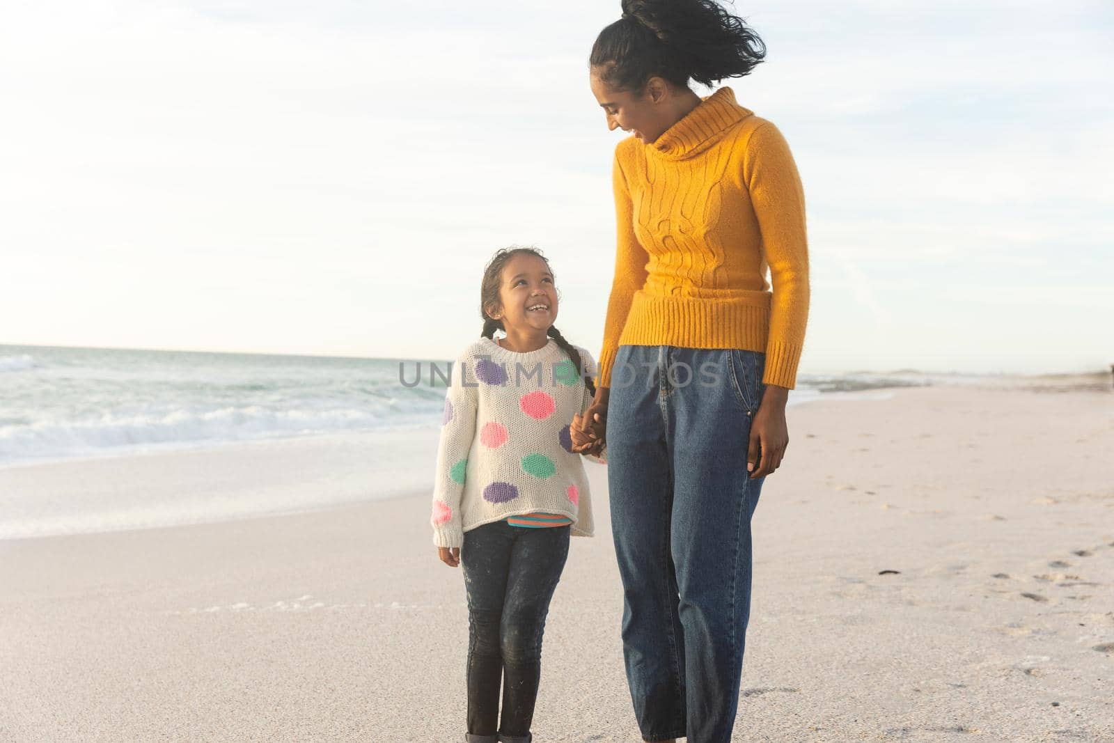 Happy biracial mother and daughter holding hands while looking at each other on beach against sky by Wavebreakmedia