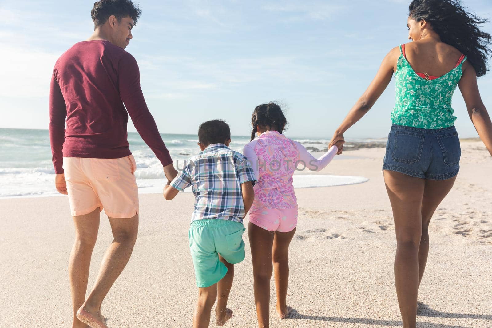 Rear view of biracial parents holding hands with children walking on sand at beach during sunny day. family, lifestyle and weekend.