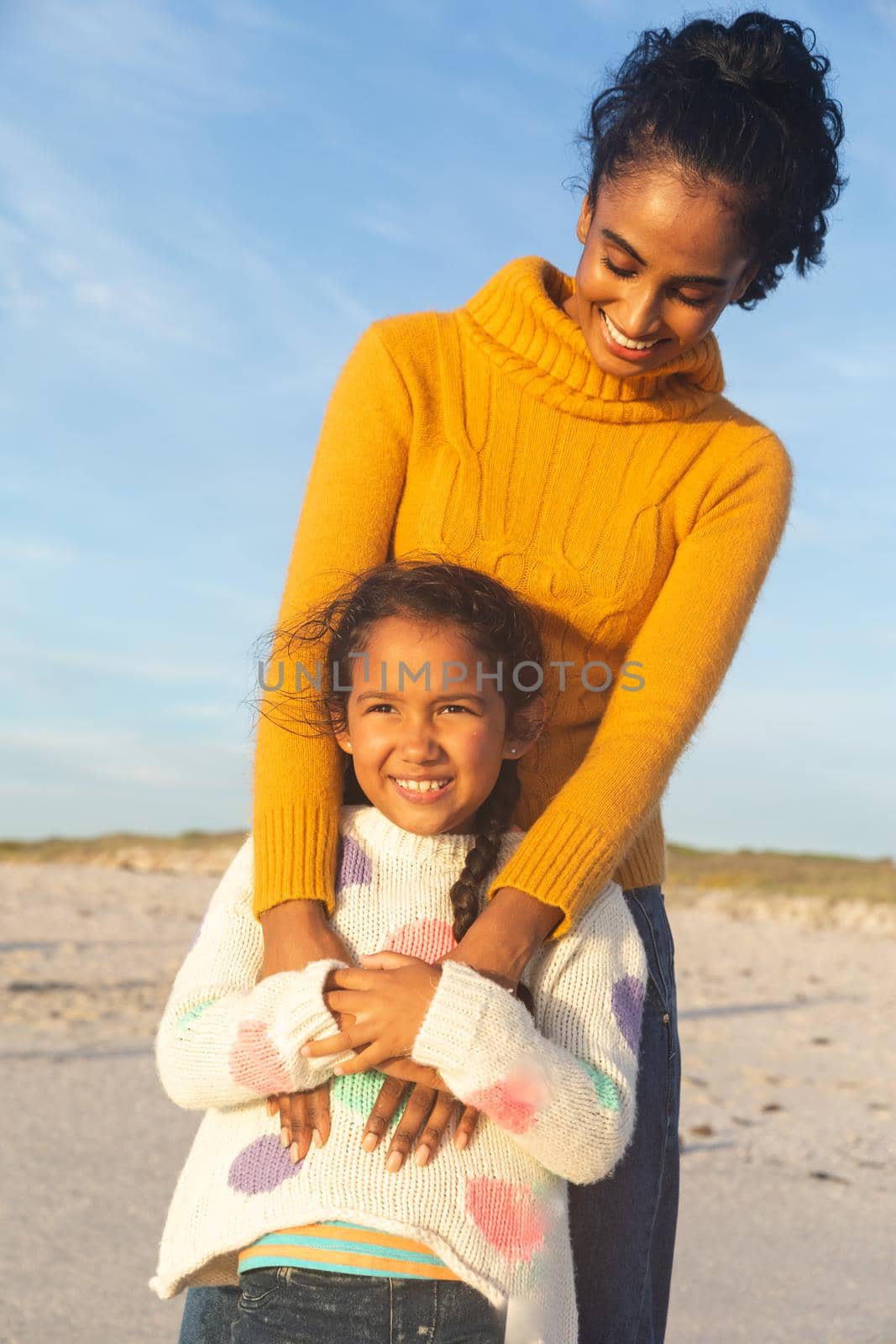 Happy young biracial woman embracing daughter from behind while standing at beach against sky. family, lifestyle and weekend.