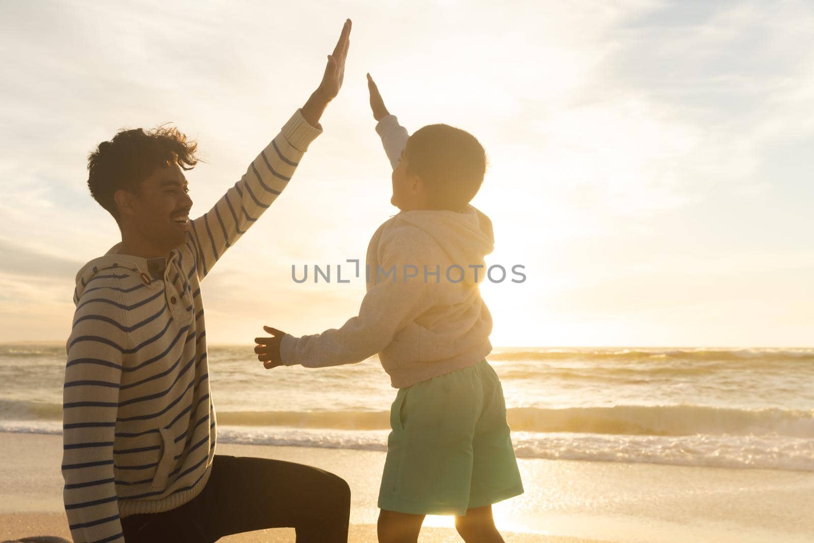 Biracial boy giving high-five to smiling father kneeling at beach against sky during sunset. family, lifestyle and weekend.