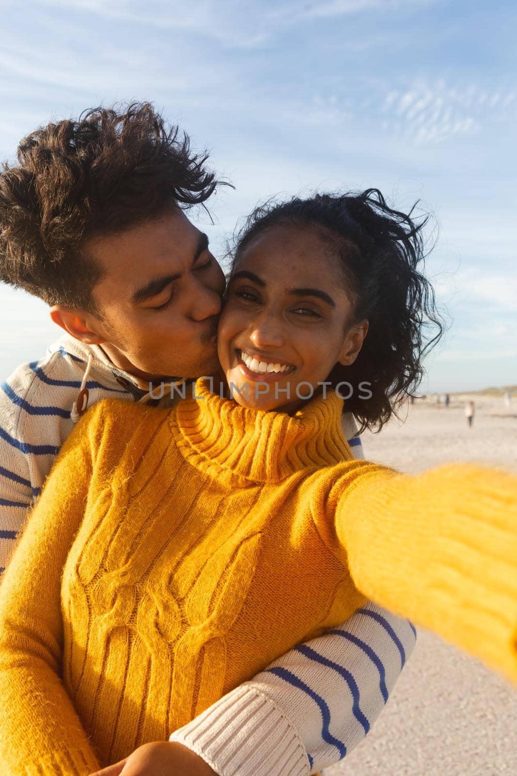 Portrait of smiling biracial woman taking selfie of boyfriend hugging and kissing on cheek at beach by Wavebreakmedia