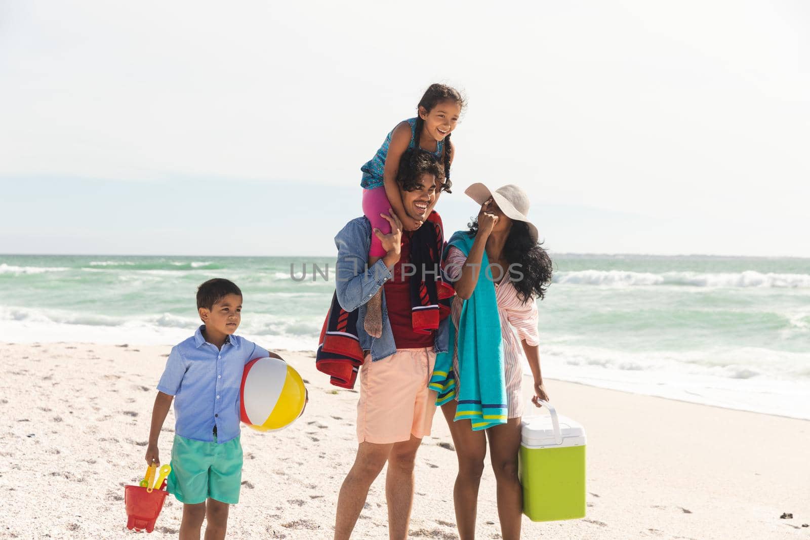 Happy biracial man carrying daughter on shoulders while walking with woman and son at beach. family, lifestyle and weekend.