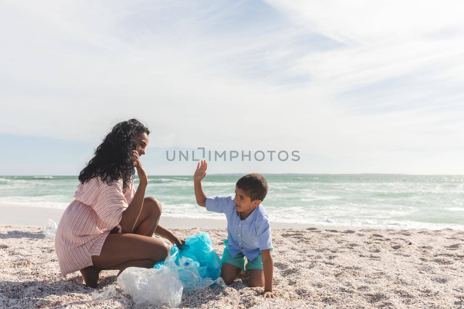 Happy biracial mother and son giving high-five while collecting garbage in bag at beach against sky by Wavebreakmedia