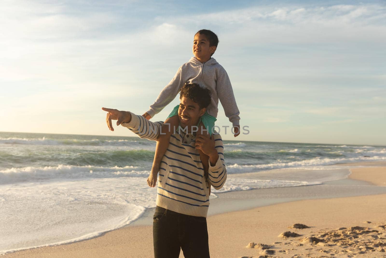 Happy biracial father pointing to son while carrying him on shoulders at beach against sky. family, lifestyle and weekend.
