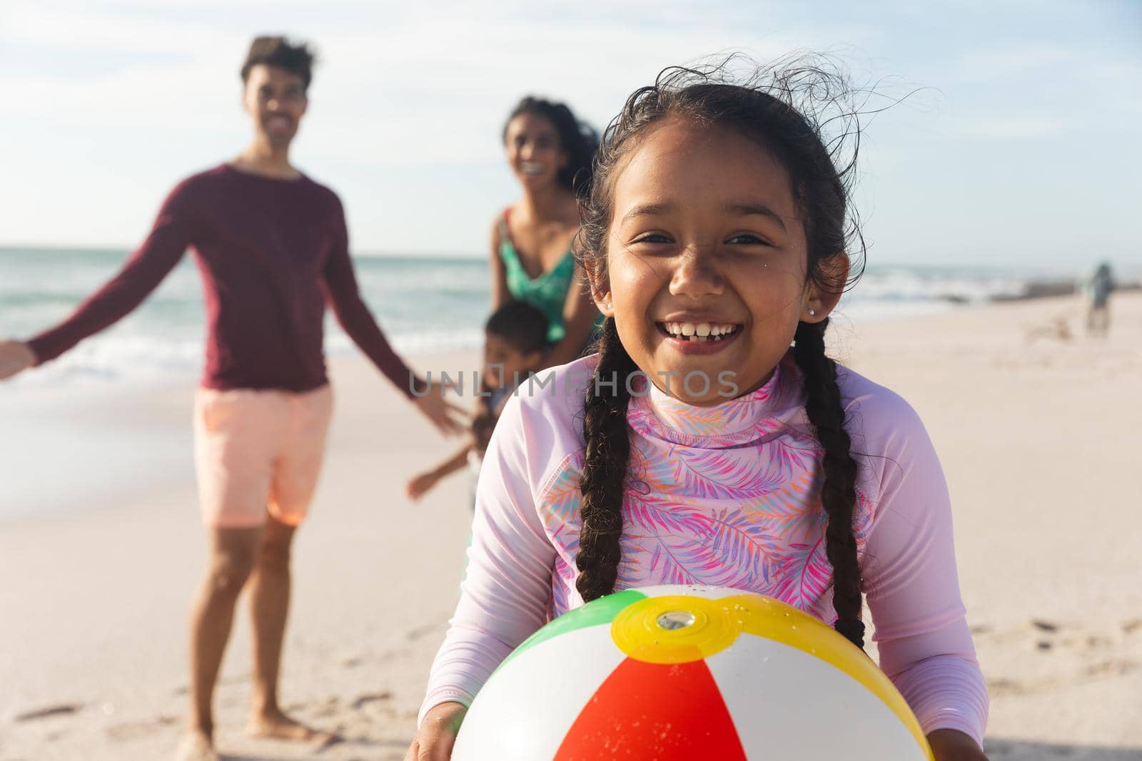 Portrait of smiling biracial girl holding ball with family at beach on sunny day by Wavebreakmedia