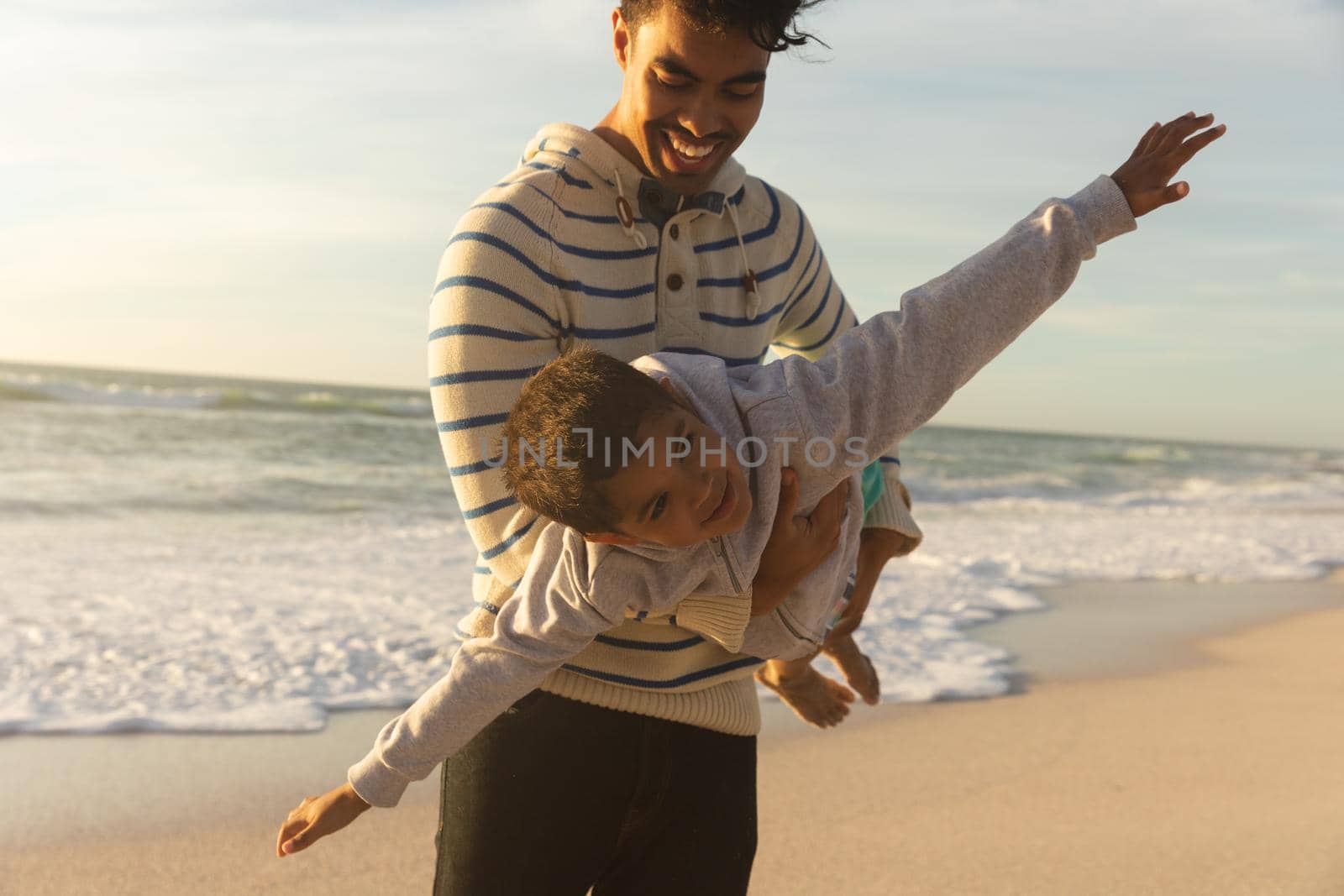 Happy biracial man carrying son pretending to fly with arms outstretched at beach during sunset. family, lifestyle and weekend.