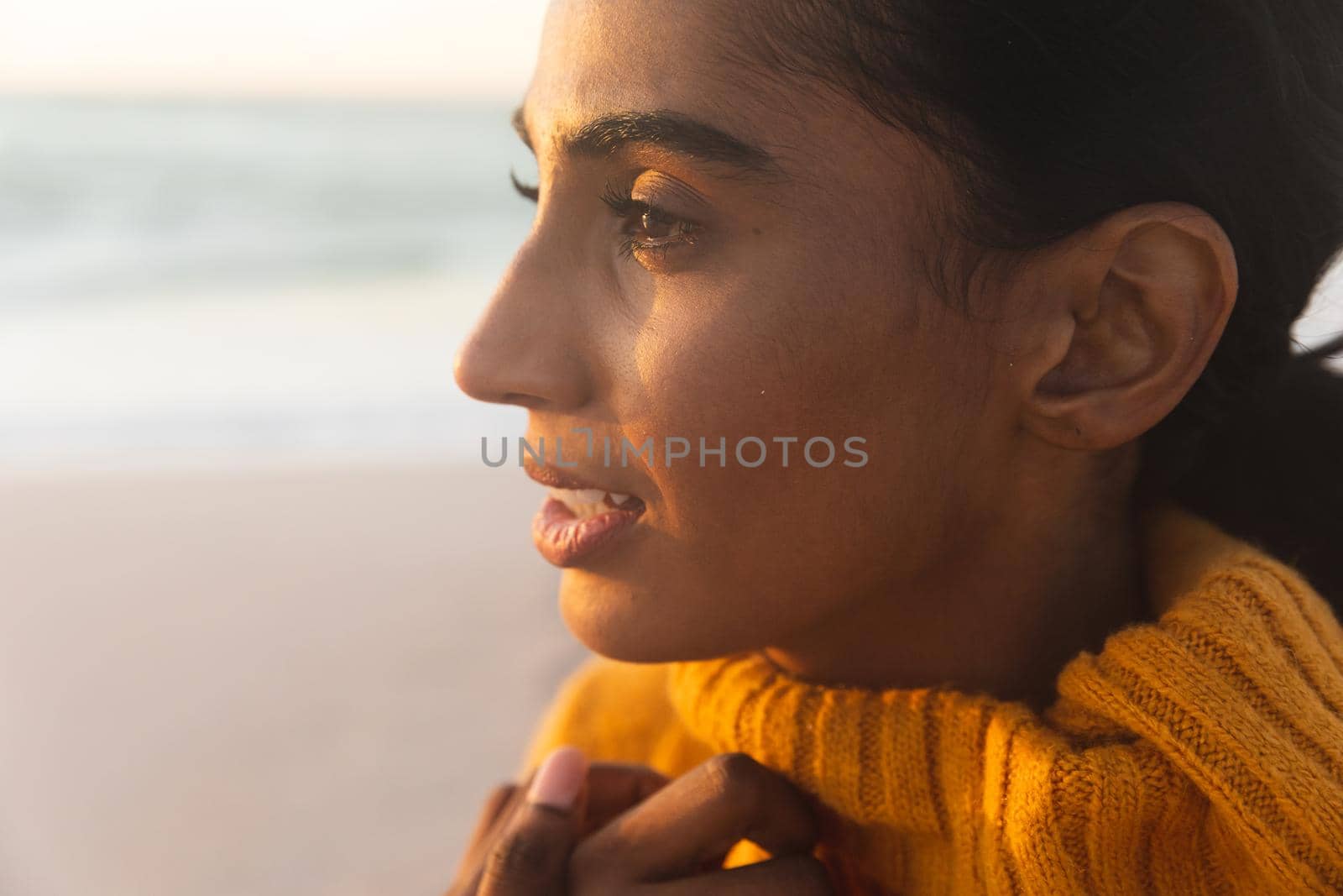 Close-up of young thoughtful biracial woman in sweater looking away at beach during sunset by Wavebreakmedia