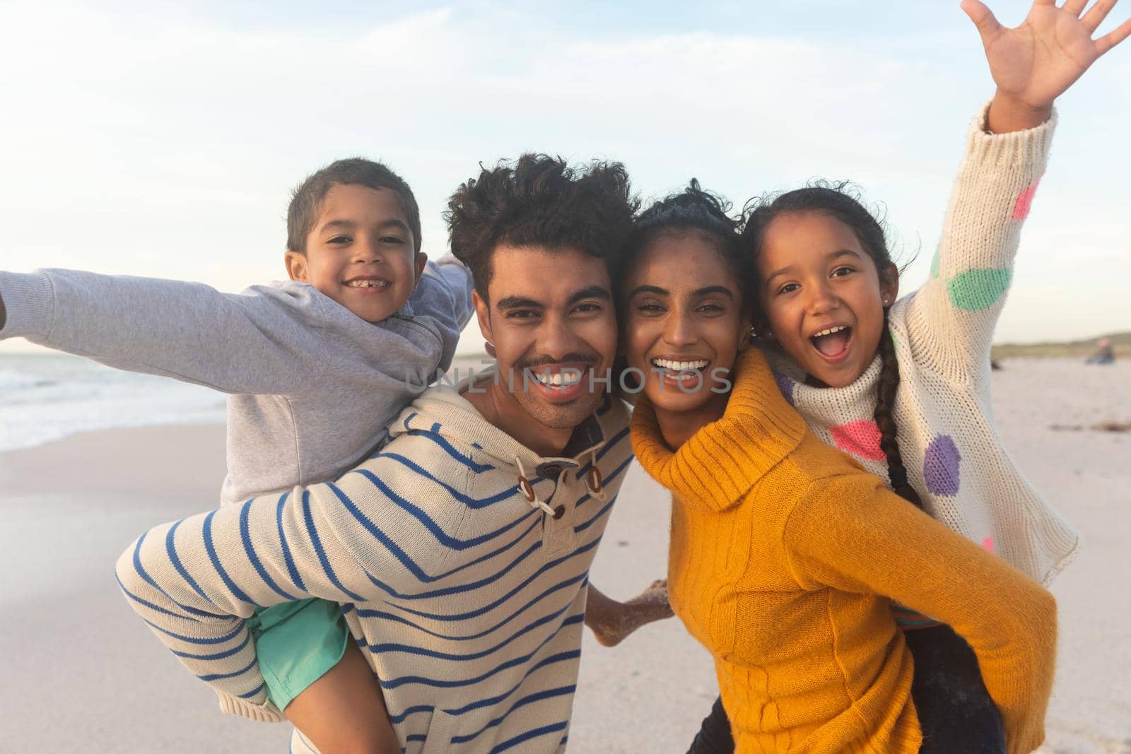 Portrait of happy biracial father and mother giving piggyback rides to kids at beach during sunset. family, lifestyle and weekend.