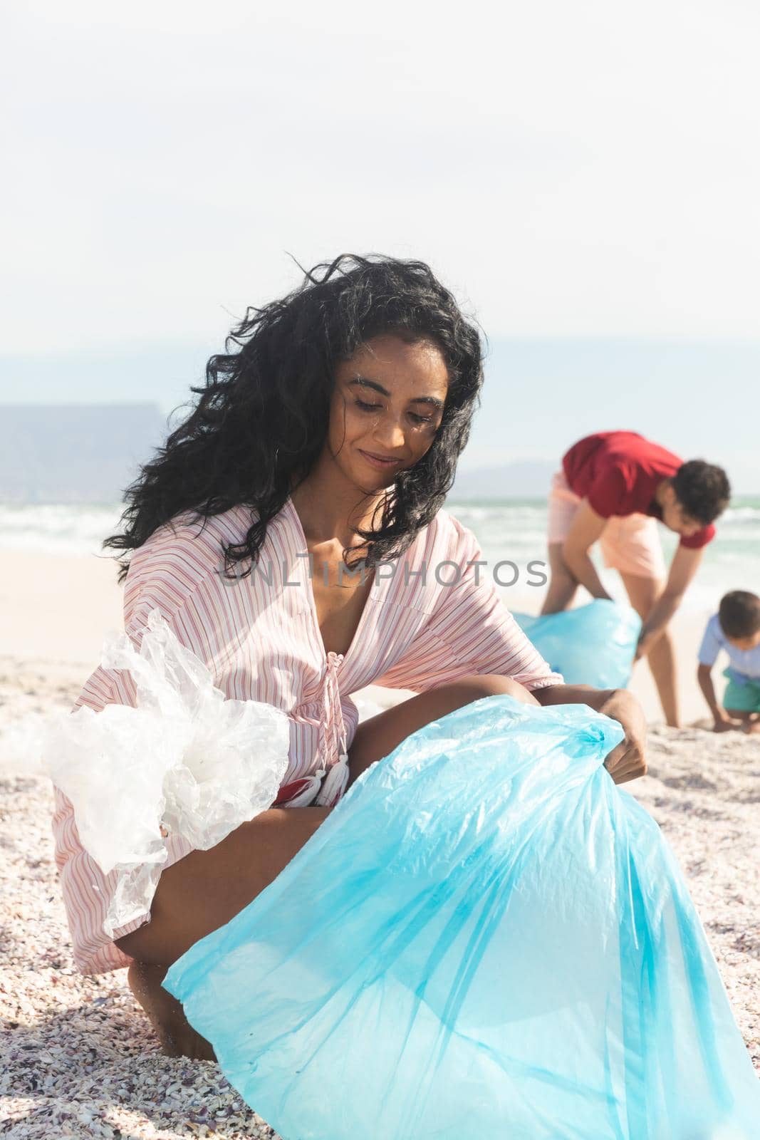 Smiling biracial woman collecting waste in blue plastic bag with family at beach by Wavebreakmedia