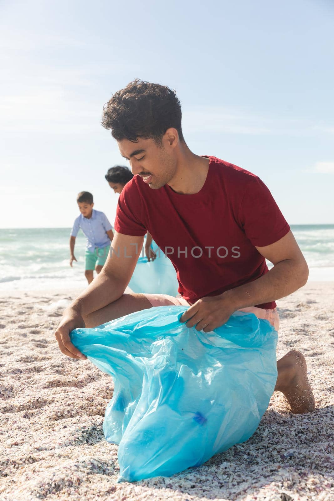 Biracial man kneeling while collecting garbage in blue plastic bag against sky with copy space by Wavebreakmedia