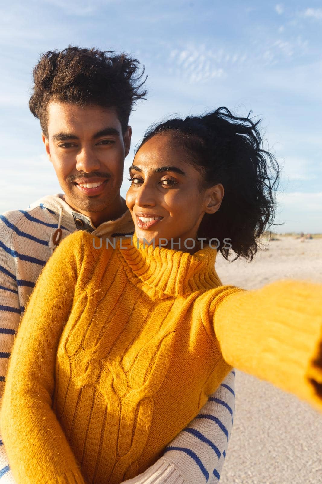 Portrait of smiling young beautiful biracial woman taking selfie with boyfriend at beach against sky by Wavebreakmedia