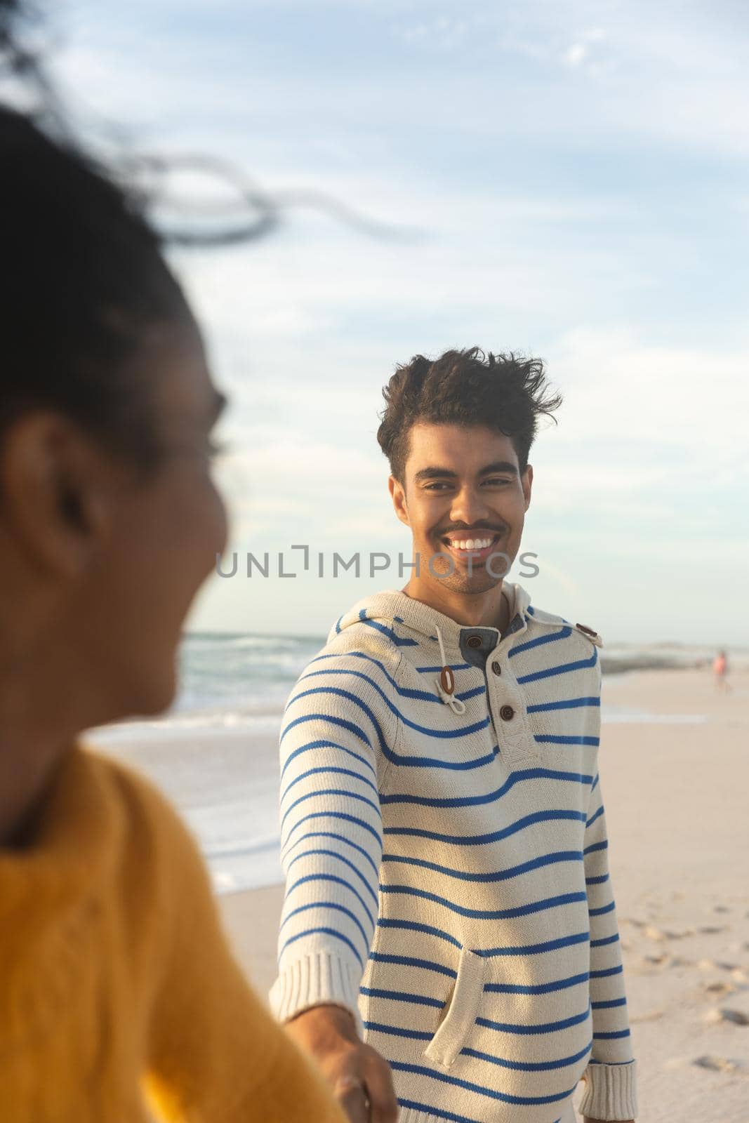 Smiling biracial man holding hand of girlfriend while standing behind her at beach against sky by Wavebreakmedia