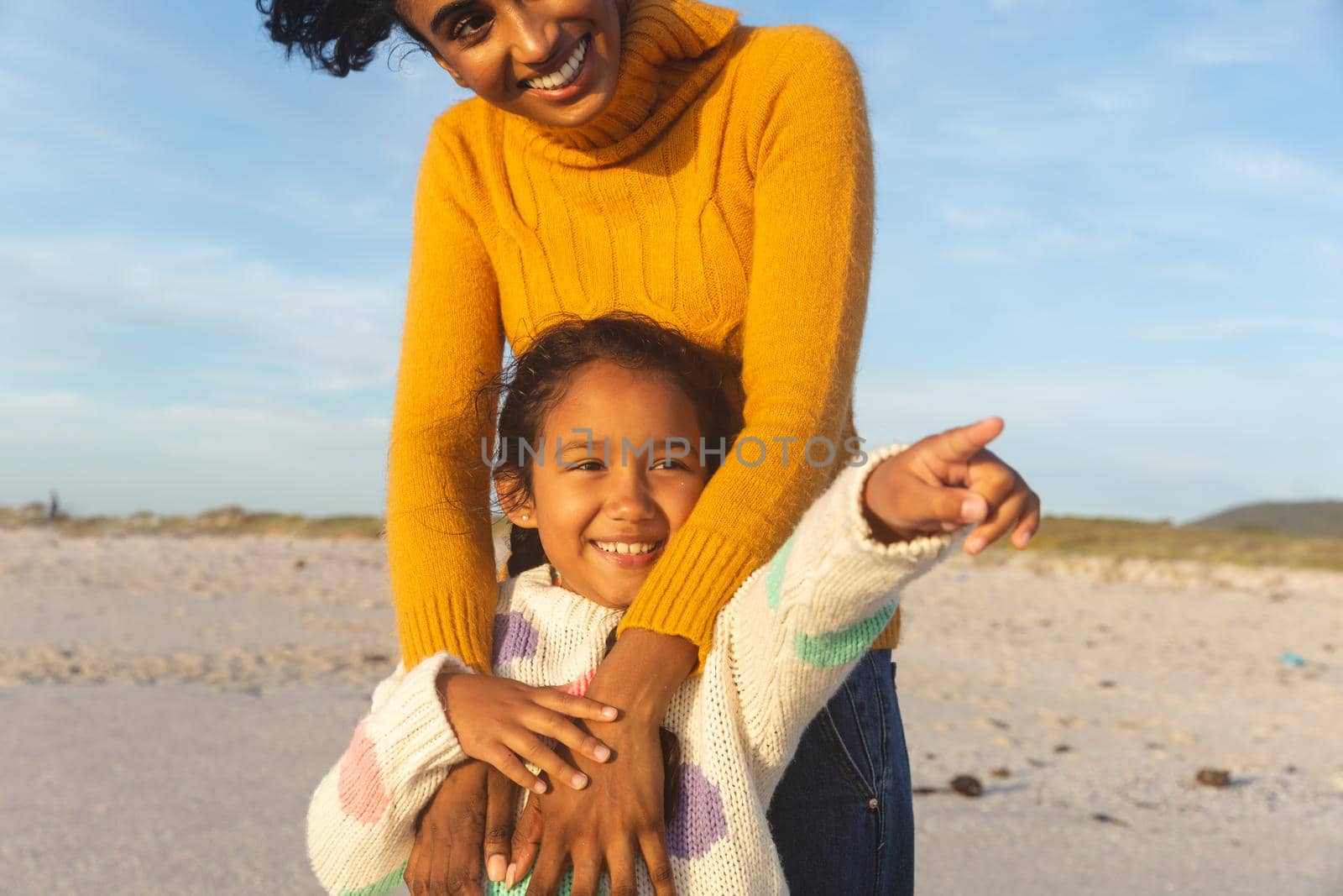 Happy biracial girl pointing to mother standing behind her at beach against sky. family, lifestyle and weekend.