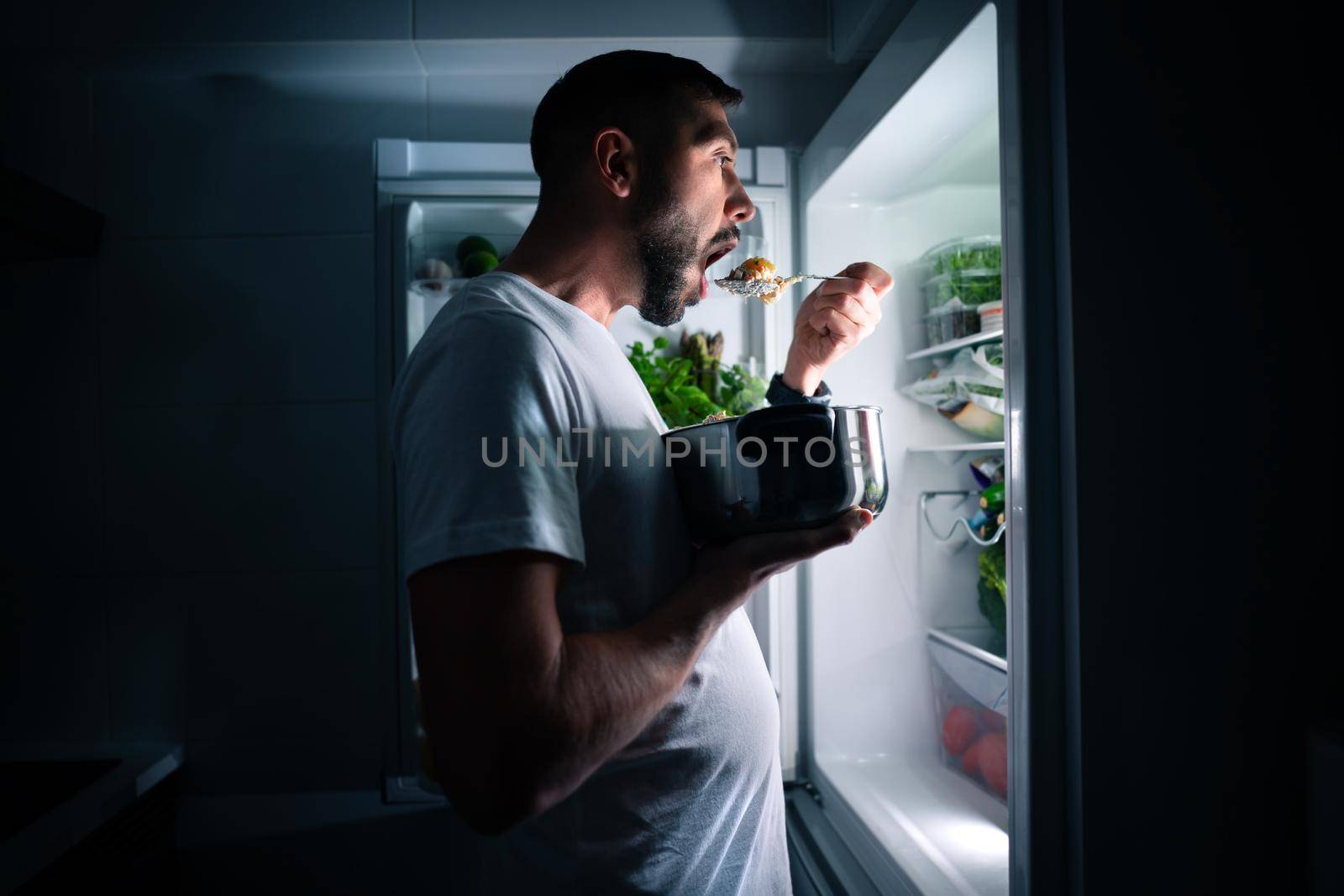 Hungry man eating food at night from open fridge. Man taking midnight snack from refrigerator. High quality photo