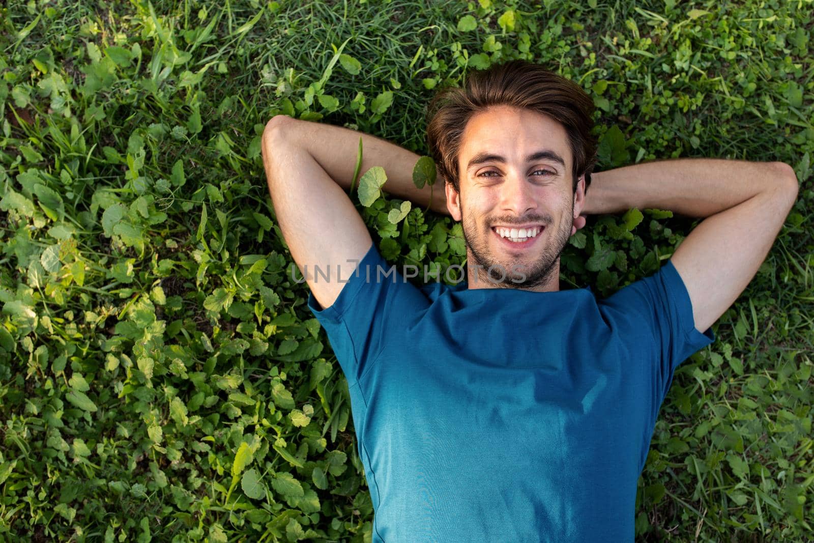Top view of happy smiling young man relaxing lying on grass looking at camera. Copy space. by Hoverstock