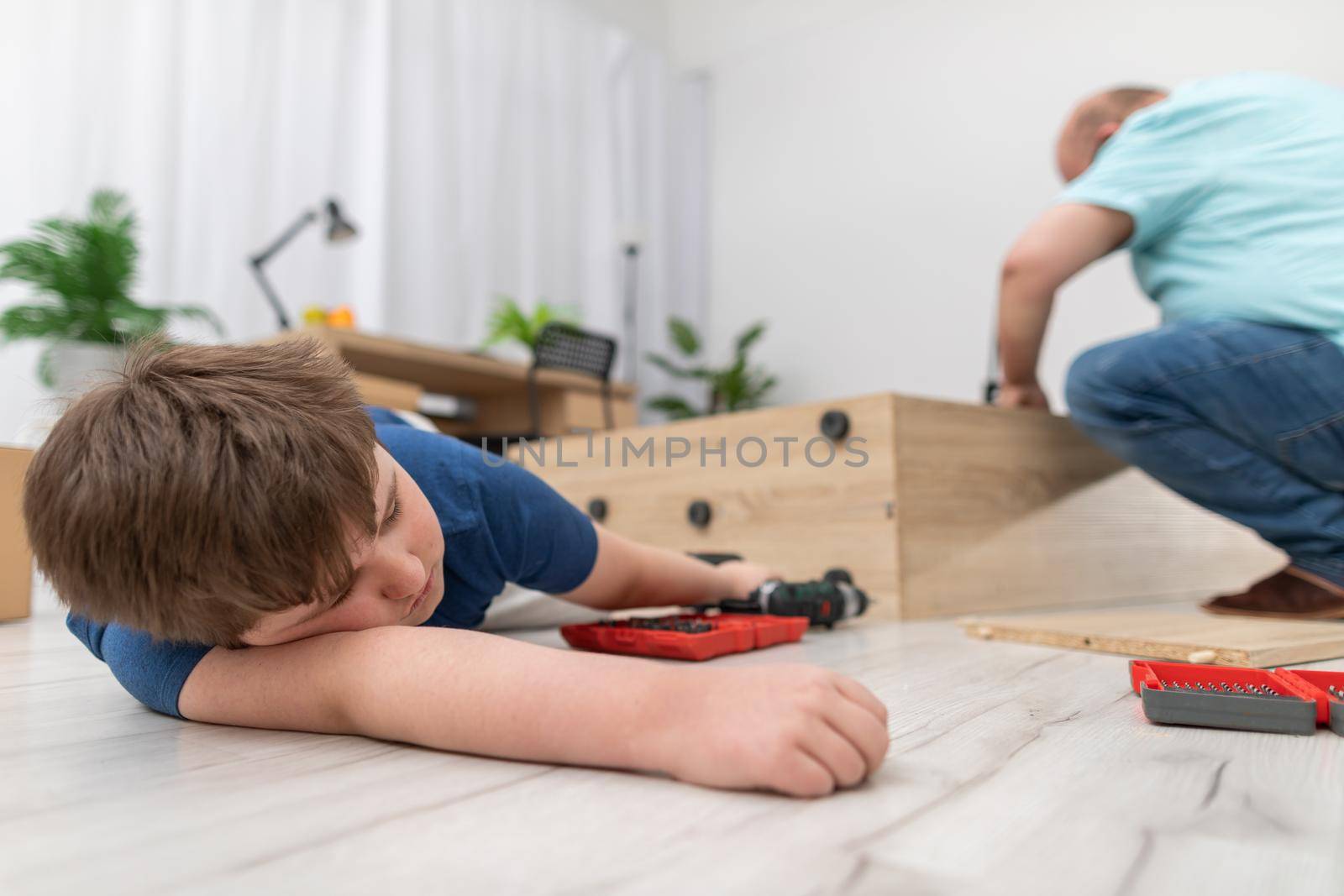 Dad is twisting furniture in the living room and son has fallen asleep on the floor. by fotodrobik